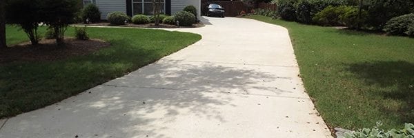 A concrete driveway curves slightly to the right leading up to a garage attached to a house. The driveway is flanked by well-kept green grass and shrubs. Shadows of trees are cast on the driveway. The house has light-colored siding, and a car is parked in the driveway.