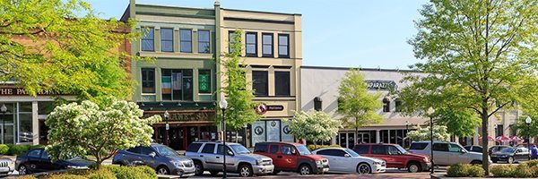 A quaint downtown street scene showing a row of various colorful commercial buildings including shops and businesses. Several cars are parked along the street, and trees with green foliage line the road, adding a touch of nature to the urban setting.