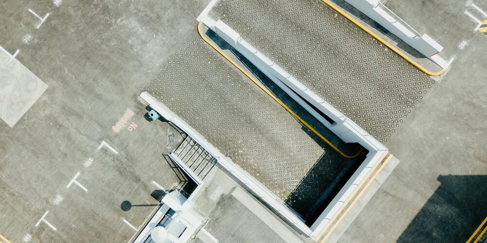 Aerial view of a multi-level parking structure with beige walls and flat grey surfaces. The image captures the upper levels, showing a ramp leading to a higher floor and a section marked with "No Parking" in faded red letters. Shadows fall across parts of the structure.